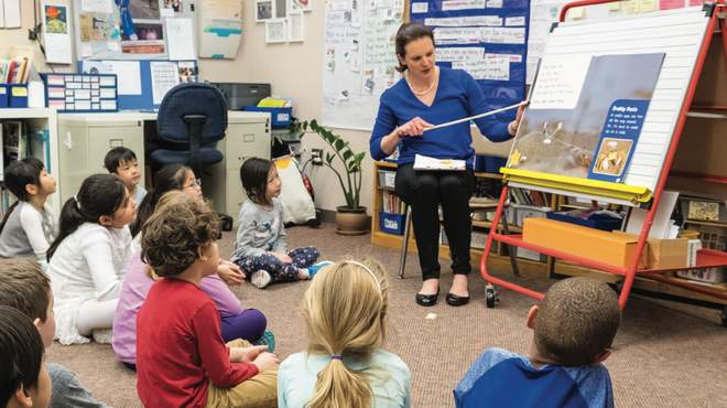 Photo of teacher pointing at large book on an easel while students look on while seated on the floor